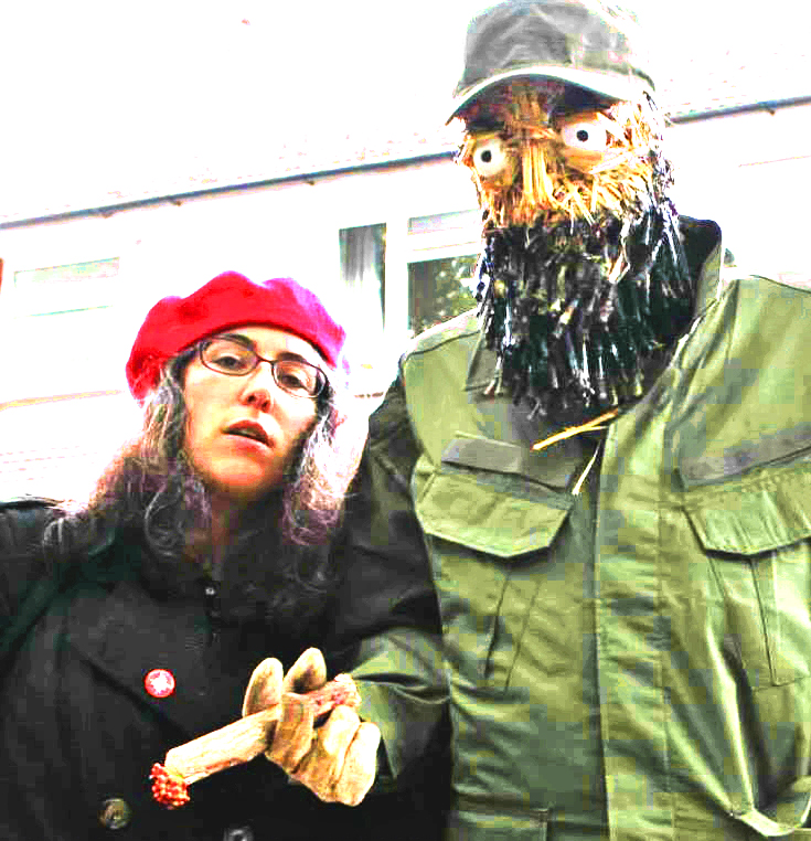 A white woman in her younger days with long dark hair, black jacket and a red beret, leaning in with a tall scarecrow Fidel Castro, as both stare intensely into the lens